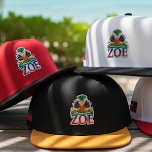 May include: A collection of flat-brimmed baseball caps in red, black, and white, with a gold or black brim. Each cap features a colorful emblem with the word "ZOE" and a Haitian coat of arms. The caps are displayed on a wooden surface.