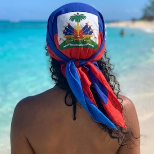 May include: A blue, red, and white headscarf featuring the Haitian coat of arms. The scarf is tied at the back of the head, with the colors of the Haitian flag prominently displayed. The person is on a beach with turquoise water.