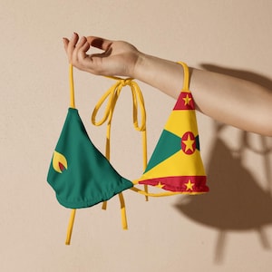 Peut inclure: Haut de bikini à deux pièces avec le drapeau de la Grenade. Le haut de bikini est tenu par une main. Le haut a un triangle vert avec un croissant jaune et un triangle jaune avec des étoiles rouges et une étoile centrale.