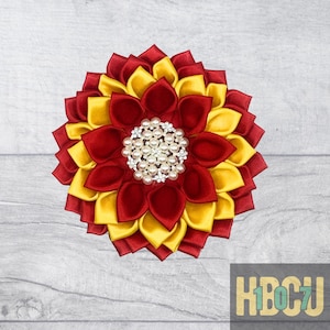 May include: A handmade, layered fabric flower in red and yellow, with a pearl and rhinestone center. The flower is a circular design, with the petals arranged in a radial pattern. The flower is a decorative accessory.