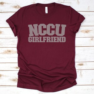 K&ouml;nnte beinhalten: Burgunderfarbenes T-Shirt mit dem Text "NCCU GIRLFRIEND" in grauen Buchstaben.