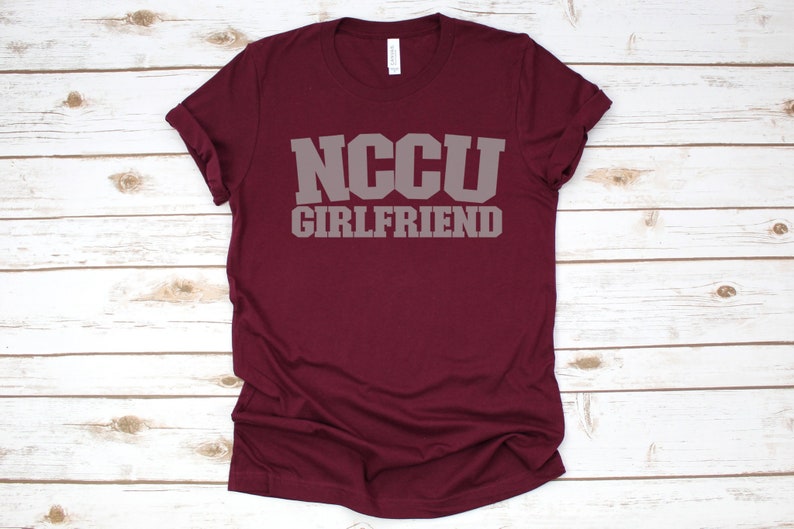 K&ouml;nnte beinhalten: Burgunderfarbenes T-Shirt mit dem Text "NCCU GIRLFRIEND" in grauen Buchstaben.