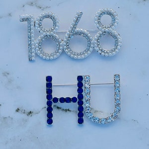 May include: Two decorative brooches. One brooch spells out "1868" with faux pearls and clear rhinestones. The other brooch spells "HU" with blue and clear rhinestones.