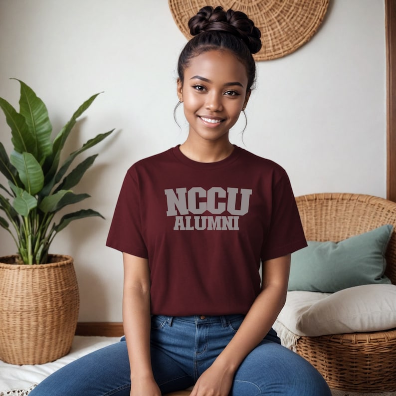 K&ouml;nnte beinhalten: Ein maroonfarbenes T-Shirt mit dem Text "NCCU ALUMNI" in wei&szlig;en Buchstaben.