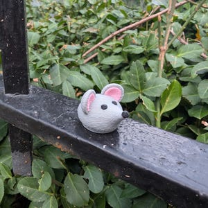 Op de afbeelding: Een kleine, grijze muizenfiguur met roze gevoerde oren en een zwarte neus zit op een zwarte metalen staaf. De muis is rond en glad, in contrast met de ruwe textuur van de staaf. Groen gebladerte vult de achtergrond.