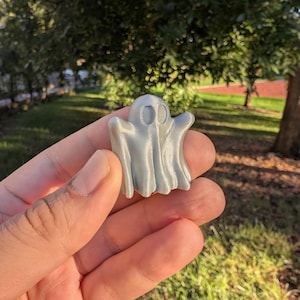 May include: A small, light gray ghost figurine with a friendly face. The ghost has a draped sheet design and is held in a hand against a background of green grass and trees. The figurine is likely a Halloween or spooky-themed decoration.