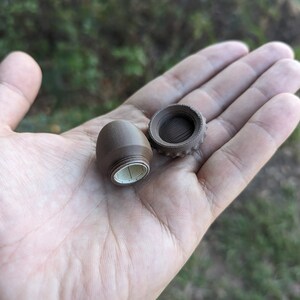 May include: Two brown, plastic, cylindrical containers with lids. One container is open and the other is closed. The open container has a white, paper label with black markings.