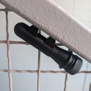May include: A matte black cylindrical device featuring a textured screw-on cap at one end and two protruding rectangular mounting brackets along its side. The object has a smooth, uniform finish and is positioned diagonally against a light-coloured surface.