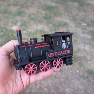 May include: Black toy train with red detailing, held in a hand. The train features red wheels and the number 1. The text "GEOCACHE" is printed on the side. This is a geocaching container, designed to look like a miniature train.