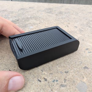 May include: A matte black rectangular storage box featuring a sliding tambour door mechanism on the top surface. The container has a smooth, solid finish with a small, elongated handle integrated into the ribbed sliding panel for easy opening.