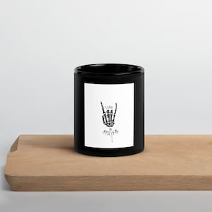 Könnte beinhalten: Schwarze Keramiktasse mit einem weißen Design, das eine Skeletthand zeigt, die ein Rock'n'Roll-Zeichen macht. Der Text lautet "Ich liebe mich selbst & ich".