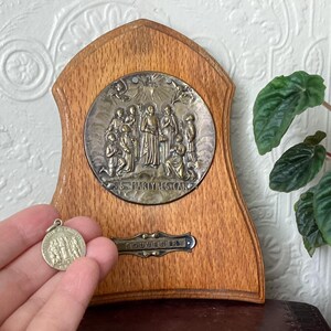 May include: A wooden plaque with a metal medallion depicting figures and the text "S. MARTYRES CAN". A small, round pendant with similar imagery is held nearby. A metal plate below the medallion reads "SOUVENIR".