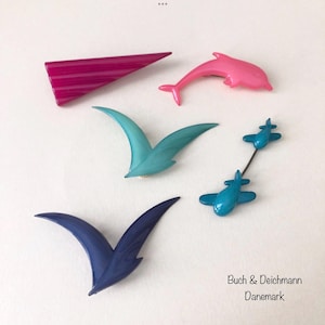 May include: Five vintage plastic brooches in various colors and shapes, including a pink dolphin, a teal seagull, a blue seagull, a blue airplane, and a pink triangle. The brooches are arranged on a white background. The text "Buch & Deichmann Danmark" is visible in the bottom right corner.