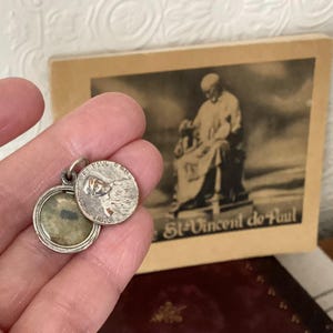 May include: A silver locket pendant with an open circular charm. The charm features an embossed image of a saint and the words "St. Paul" and "Paris". A sepia-toned image of a saint is in the background.