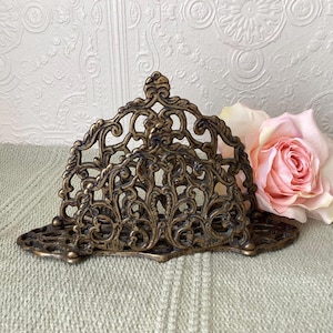 Peut inclure: Porte-serviettes en métal de couleur laiton antique avec un design filigrané orné. Le support a un dessus incurvé et une base rectangulaire, avec des volutes et des motifs floraux complexes. Une rose rose est visible en arrière-plan.