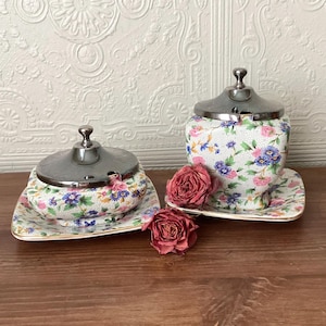 May include: Two ceramic containers with silver-toned metal lids and matching saucers, featuring a white base with a dense pattern of small pink, blue, and yellow flowers and green leaves, accompanied by two dried red roses placed in the centre.