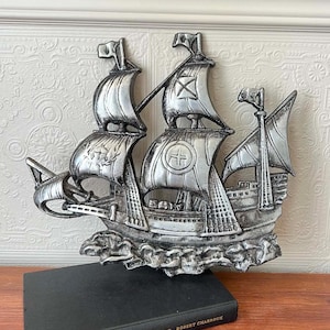 May include: A silver-coloured wall plaque shaped like a three-masted sailing ship. The ship features detailed sails with embossed markings, including a cross and a circular emblem, set against a textured base representing waves in a metallic finish.