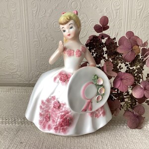 May include: A vintage porcelain figurine of a woman in a white dress with pink floral accents. She holds a matching hat with pink ribbon and floral embellishments. The figurine has blonde hair and a pink bow. Decorative dried flowers are in the background.