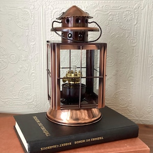 Peut inclure: Une lanterne de couleur cuivre avec des panneaux de verre et une poignée. Elle est posée sur un livre noir avec des lettres dorées. Le livre est intitulé "TRESORS DU MONDE ROBERT CHARROUX". La lanterne a un dessus décoratif.