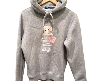May include: A gray hooded sweatshirt featuring a colorful cartoon bear graphic holding a basketball. The bear is wearing a striped shirt and sneakers. The text "POLO BEAR by RALPH LAUREN" is printed below the bear.