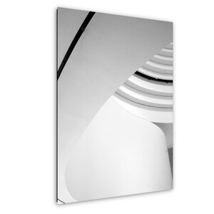 May include: A black and white photograph of a spiral staircase interior. The image features curved white architectural walls and a dark diagonal line cutting across the upper left corner, creating a high-contrast geometric composition.