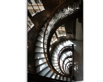 May include: An overhead view of a spiral staircase with white steps and dark wooden railings. The staircase curves downwards, with sunlight streaming through the windows, creating shadows and highlights on the steps.