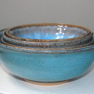 May include: A set of four ceramic bowls in varying sizes, nested together. The bowls are a vibrant turquoise blue with a speckled texture and a darker brown rim. The bowls are sitting on a white surface.
