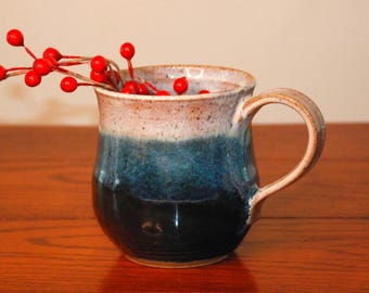 May include: A ceramic mug with a rounded body and a curved handle, featuring a glaze that transitions from a speckled cream color at the rim to a deep blue and black at the base. A sprig of small, bright red berries is placed inside the mug.