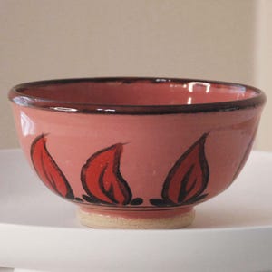 May include: A small, pink ceramic bowl with a black rim and red and black floral designs. The bowl is sitting on a white surface.
