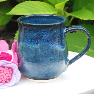 May include: A ceramic mug with a glossy, deep blue glaze that transitions from a dark navy rim to a lighter, mottled cobalt body. The mug features a curved handle and a smooth, rounded silhouette, accompanied by a cluster of pink and lavender felt flowers.