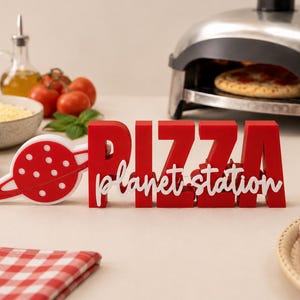 Puede incluir: Un letrero rojo y blanco con el texto PIZZA en letras de bloque en negrita y planet station en una fuente cursiva blanca. A la izquierda, se muestra un icono de planeta circular con un anillo. El letrero tiene un diseño tridimensional.
