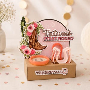 Peut inclure: Un présentoir en carton brun avec une découpe représentant une botte de cow-boy brune, des fleurs roses et un cactus. Le texte indique Tatum's FIRST RODEO et THANKS Y'ALL. Il comprend un récipient orange rond et deux emporte-pièces en fer à cheval.