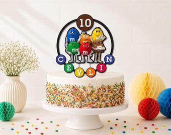 May include: A round cake topper featuring six colorful M&M characters, the number 10 in a brown circle at the top, and the name CEYLIN spelled out in individual colored circles at the bottom, all displayed on a white cake with rainbow sprinkles.