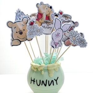 May include: A light green, round container with the word "HUNNY" in black lettering, overflowing with character cutouts on wooden sticks. The cutouts feature Winnie the Pooh and friends, with white and yellow accents.