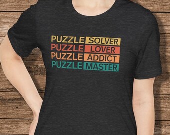 May include: Dark gray t-shirt with the words "PUZZLE SOLVER, PUZZLE LOVER, PUZZLE ADDICT, PUZZLE MASTER" in a retro font. The text is in yellow, orange, teal, and green. The shirt is a crew neck.