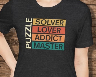 May include: A dark gray t-shirt with a vintage-style graphic that reads "PUZZLE SOLVER LOVER ADDICT MASTER". The text is in a colorful block style with a red, orange, yellow, and green color scheme.