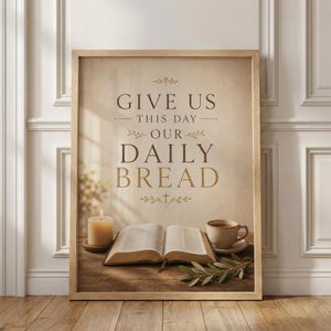 May include: Framed print with the text "GIVE US THIS DAY OUR DAILY BREAD" in brown lettering. The image includes a lit candle, an open book, a cup of coffee, and greenery, all arranged on a wooden surface.