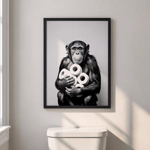 Può includere: Stampa in bianco e nero incorniciata di uno scimpanzé che tiene in mano diversi rotoli di carta igienica. L'opera d'arte è esposta sopra un gabinetto bianco in un ambiente bagno. L'immagine ha un'estetica umoristica e moderna.