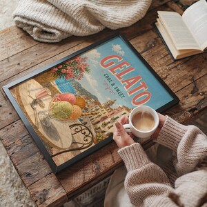 May include: A framed print with the word "GELATO" in red letters, depicting a gelato shop scene with colorful scoops of ice cream. A cup of coffee sits next to the print on a wooden table.