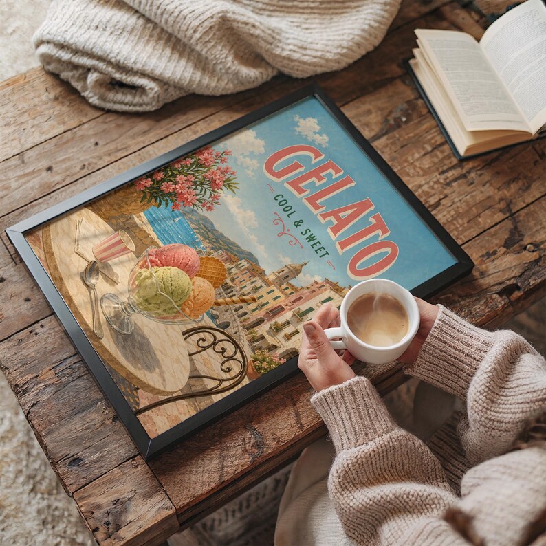 May include: A framed print with the word "GELATO" in red letters, depicting a gelato shop scene with colorful scoops of ice cream. A cup of coffee sits next to the print on a wooden table.