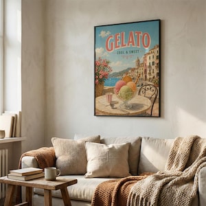 May include: A framed vintage-style poster with the word "Gelato" in pink lettering. The poster depicts a seaside scene with colorful gelato in a glass bowl on a table. A pink drink and spoon are also on the table. The poster is hanging above a sofa.