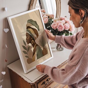 May include: A framed art print featuring an abstract illustration of a woman wearing a turban. The artwork uses a palette of earth tones, including beige, brown, and green. The woman is depicted in profile, with gold-colored earrings. The frame is white.