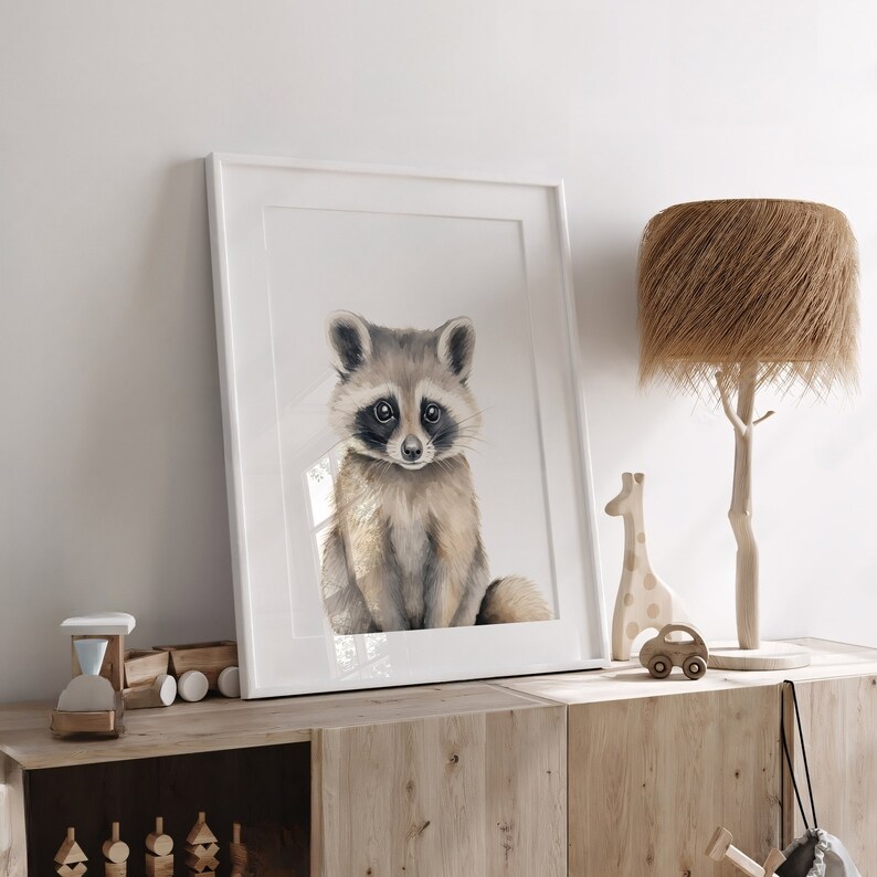 Puede incluir: Una pintura de acuarela de un mapache sentado en un marco blanco. El mapache tiene un pelaje gris y marr&oacute;n con ojos negros y una nariz negra. La pintura est&aacute; en un estante de madera con otra decoraci&oacute;n.