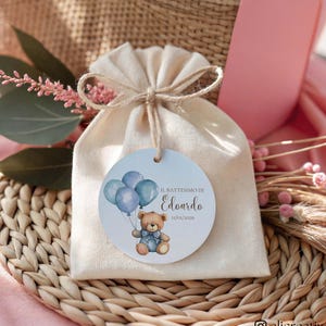 Può includere: Un piccolo sacchetto regalo in tessuto beige legato con spago, con un'etichetta rotonda con un orsetto che tiene palloncini blu. L'etichetta recita "Il Battesimo di Edoardo 11/05/2026". La borsa poggia su un tappetino intrecciato con accenti rosa e verdi.
