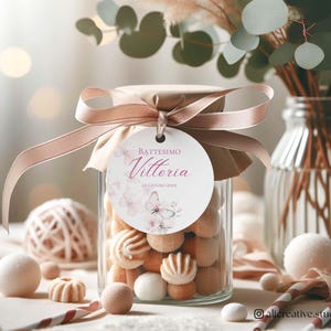 Può includere: Un barattolo di vetro pieno di caramelle assortite, legato con un nastro marrone chiaro e un'etichetta con la scritta "Battessimo Vittoria". Il barattolo è circondato da elementi decorativi, tra cui un vaso di verde e un gomitolo di spago.