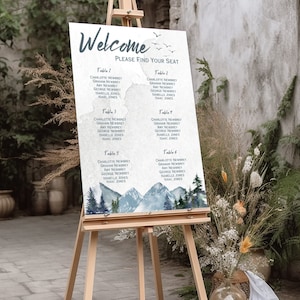 May include: A wedding seating chart with a watercolor mountain and forest design. The chart is on an easel and lists the names of guests at each table. The text reads "Welcome, Please Find Your Seat".
