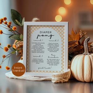 Puede incluir: Un cartel enmarcado en blanco con fondo beige y un patrón de cuadros blanco y negro. El cartel tiene el texto "DIAPER PONG" en negro, con una fuente redondeada. El cartel tiene instrucciones para un juego de tres rondas y un desempate. El cartel tiene el texto "¡buena suerte!" en la parte inferior.