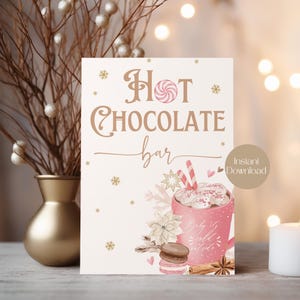 May include: Printable sign for a hot chocolate bar with a pink mug of hot chocolate with marshmallows and a candy cane, a macaron, and cinnamon sticks. The sign says "Hot Chocolate Bar" in brown text with gold snowflakes and stars.