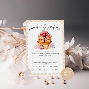 May include: A baby shower invitation with a watercolor illustration of pancakes topped with berries and syrup. The text reads "pancakes & pacifiers" and includes event details: date, time, location, and RSVP information. The design has a beige gingham border.