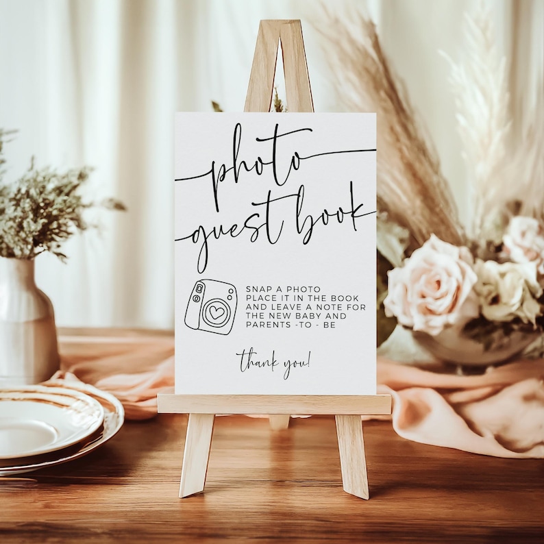 May include: A white sign on a small wooden easel with the words "photo guest book" in elegant script. Below, a camera graphic and text that reads "Snap a photo, place it in the book..." The sign is set on a wooden table.